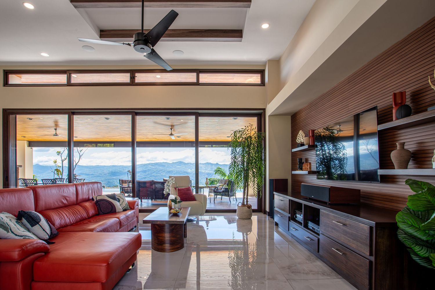 Spacious living room with red leather sofa, modern ceiling fan, and glass doors opening to a scenic view