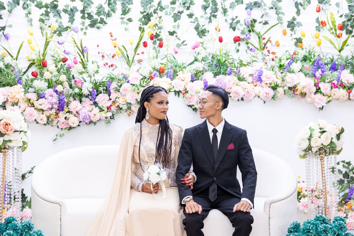 Couple sits on elegant white couch with floral arch backdrop at wedding celebration.