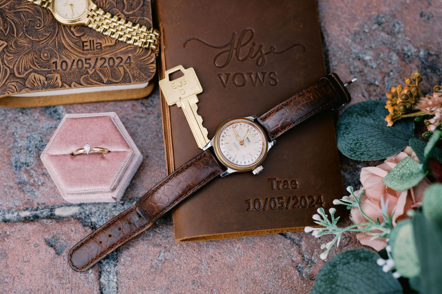 Wedding details including leather wallet, watch, pink ring box, and key on textured surface.