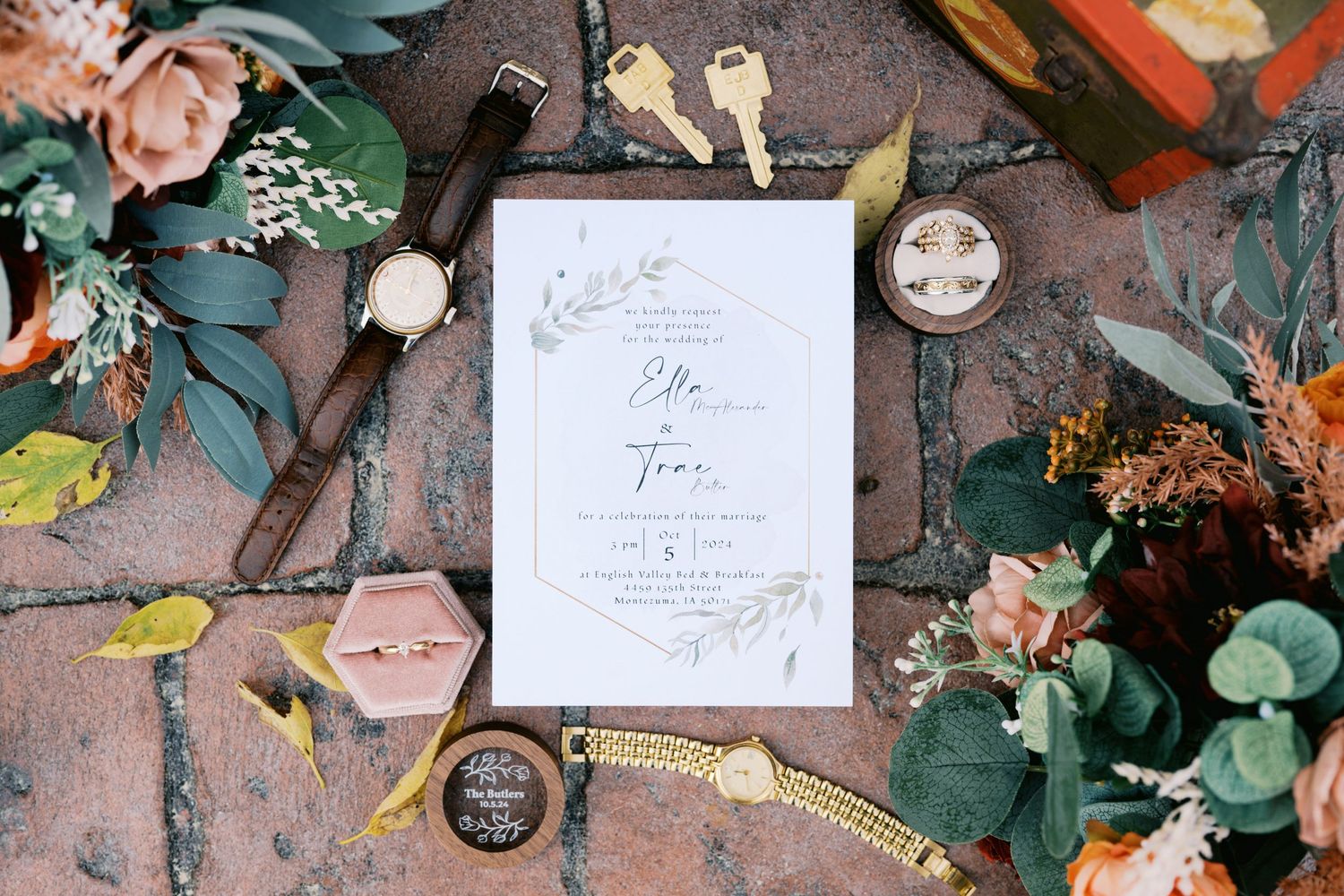 Wedding invitation and decor flat lay with vintage keys, eucalyptus leaves, flowers, and pocket watch on dark surface.