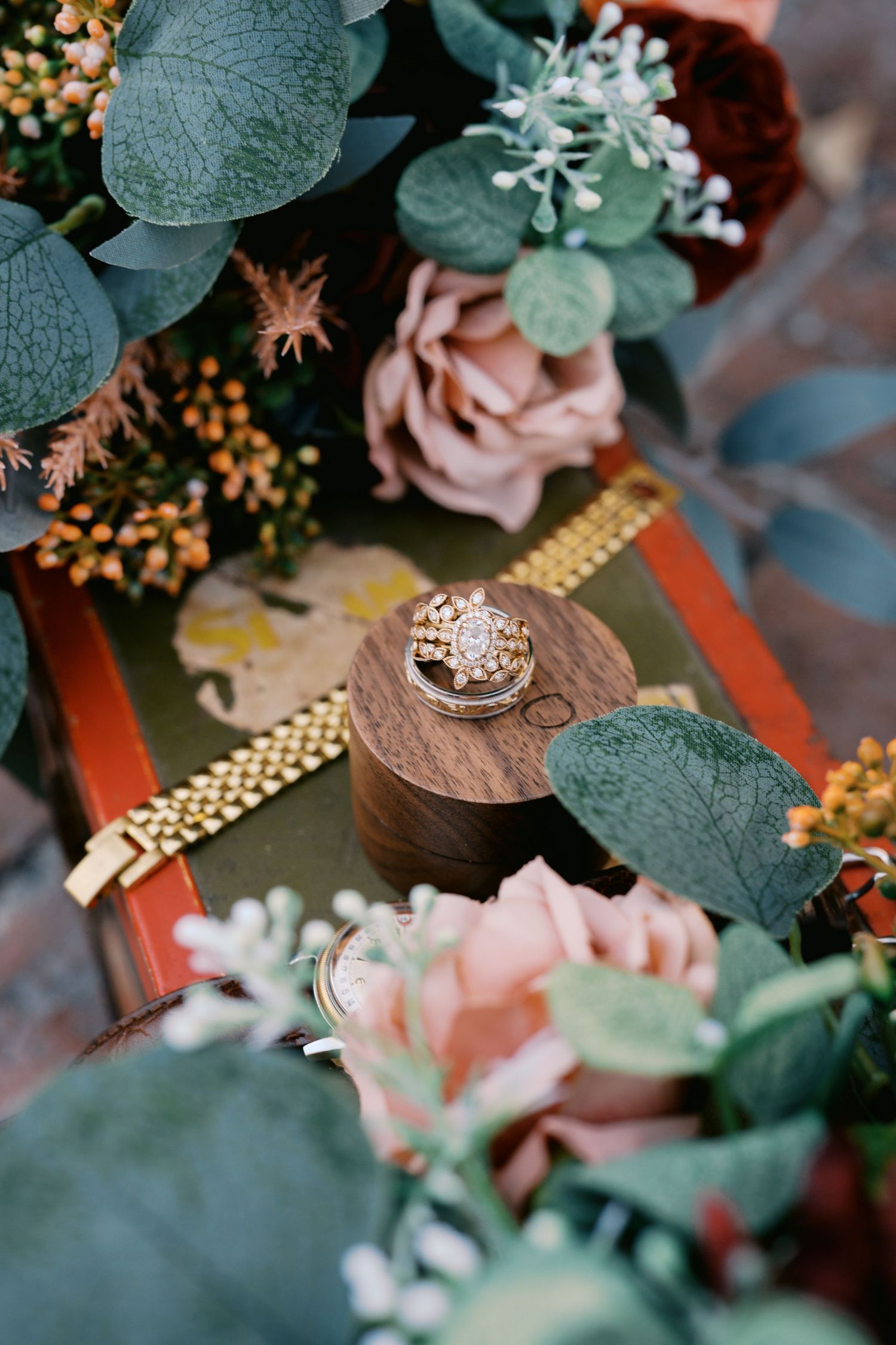 Wedding ring displayed among eucalyptus leaves and pink roses on decorative surface.