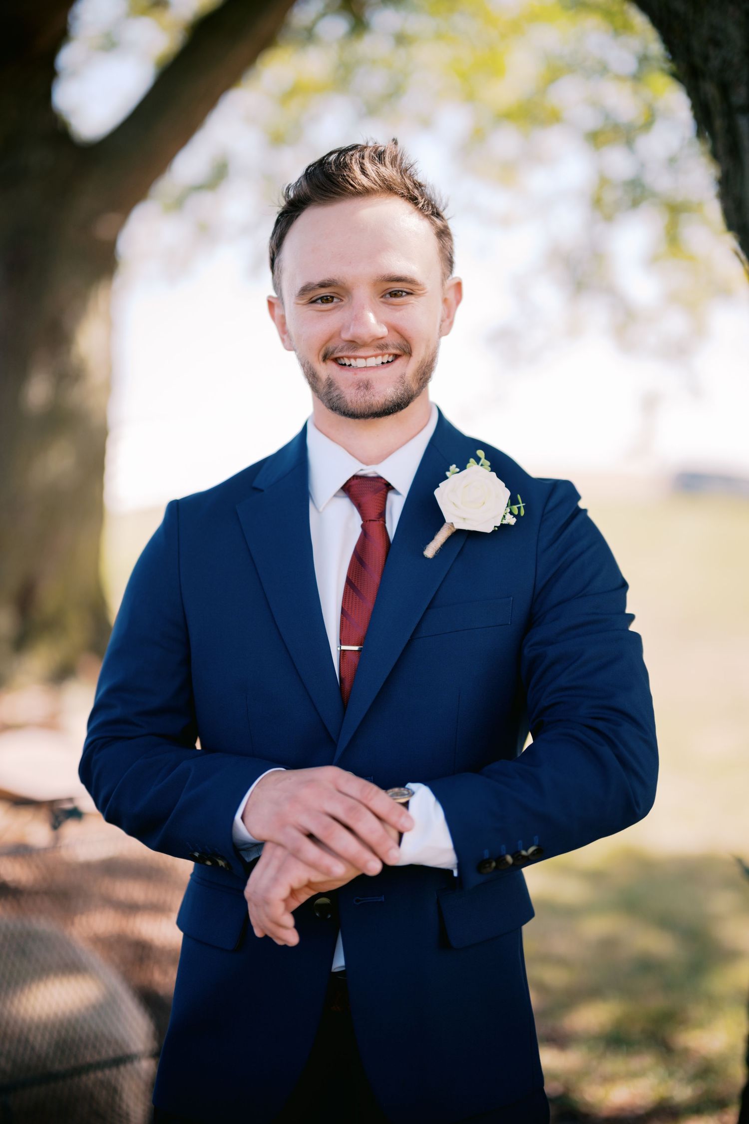 A groom in a navy suit with red tie and white boutonniere poses outdoors.