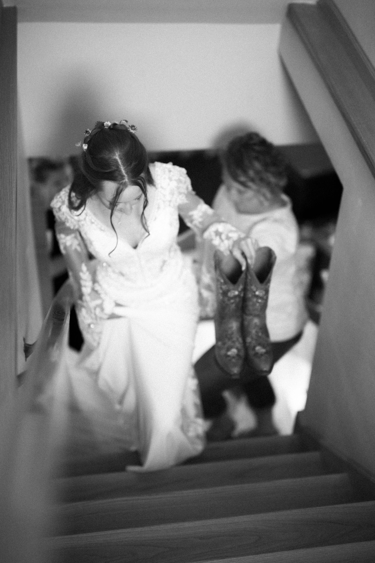 A black and white photo shows someone walking upstairs in a wedding dress while holding cowboy boots.