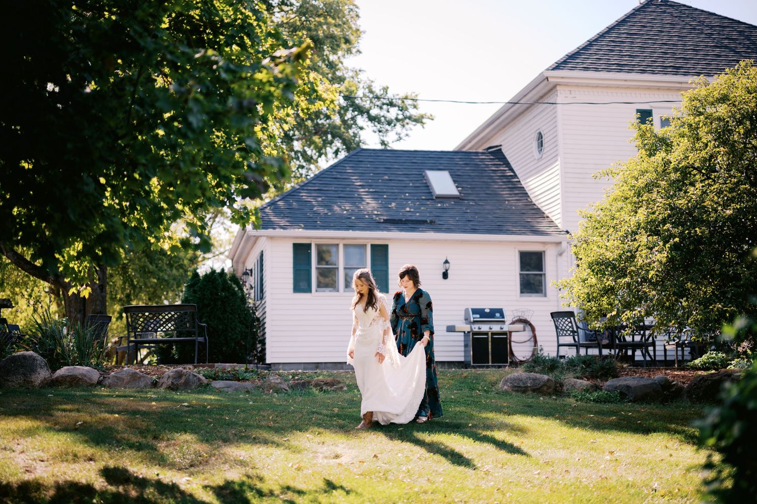Wedding couple walking across sunny lawn in front of white house with trees.