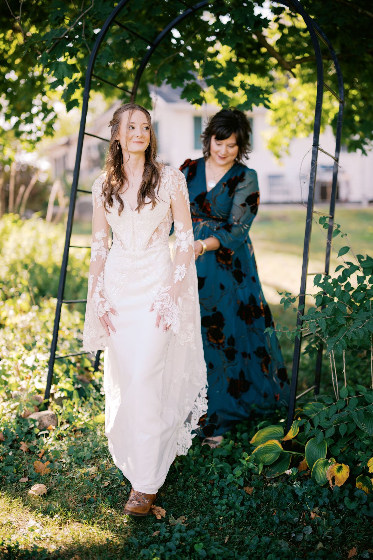A wedding dress being adjusted under a garden arch with greenery and sunlight filtering through.