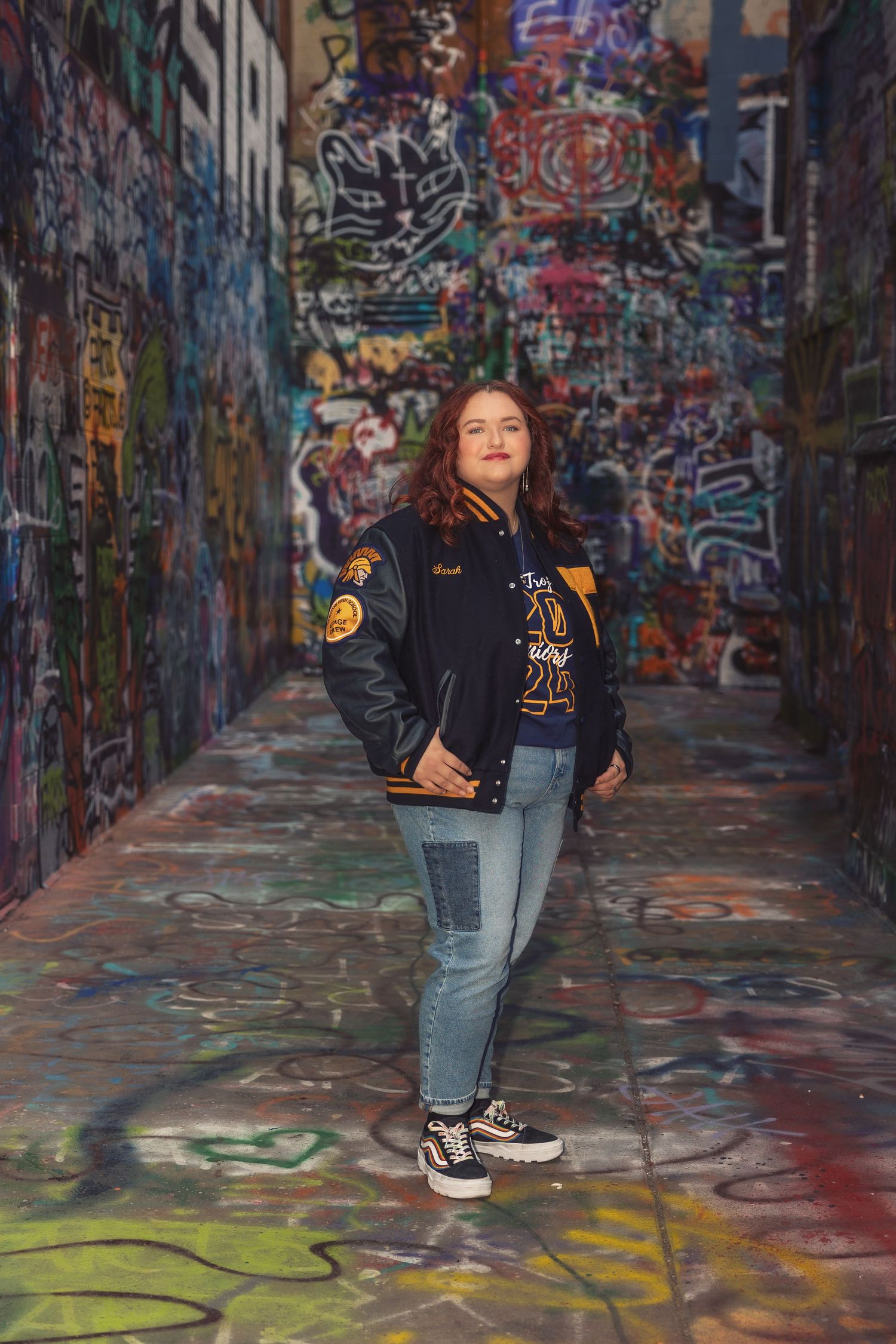 A high school senior poses casually in front of a vibrant graffiti wall wearing a varsity jacket, jeans and sneakers in Ann Arbor Michigan