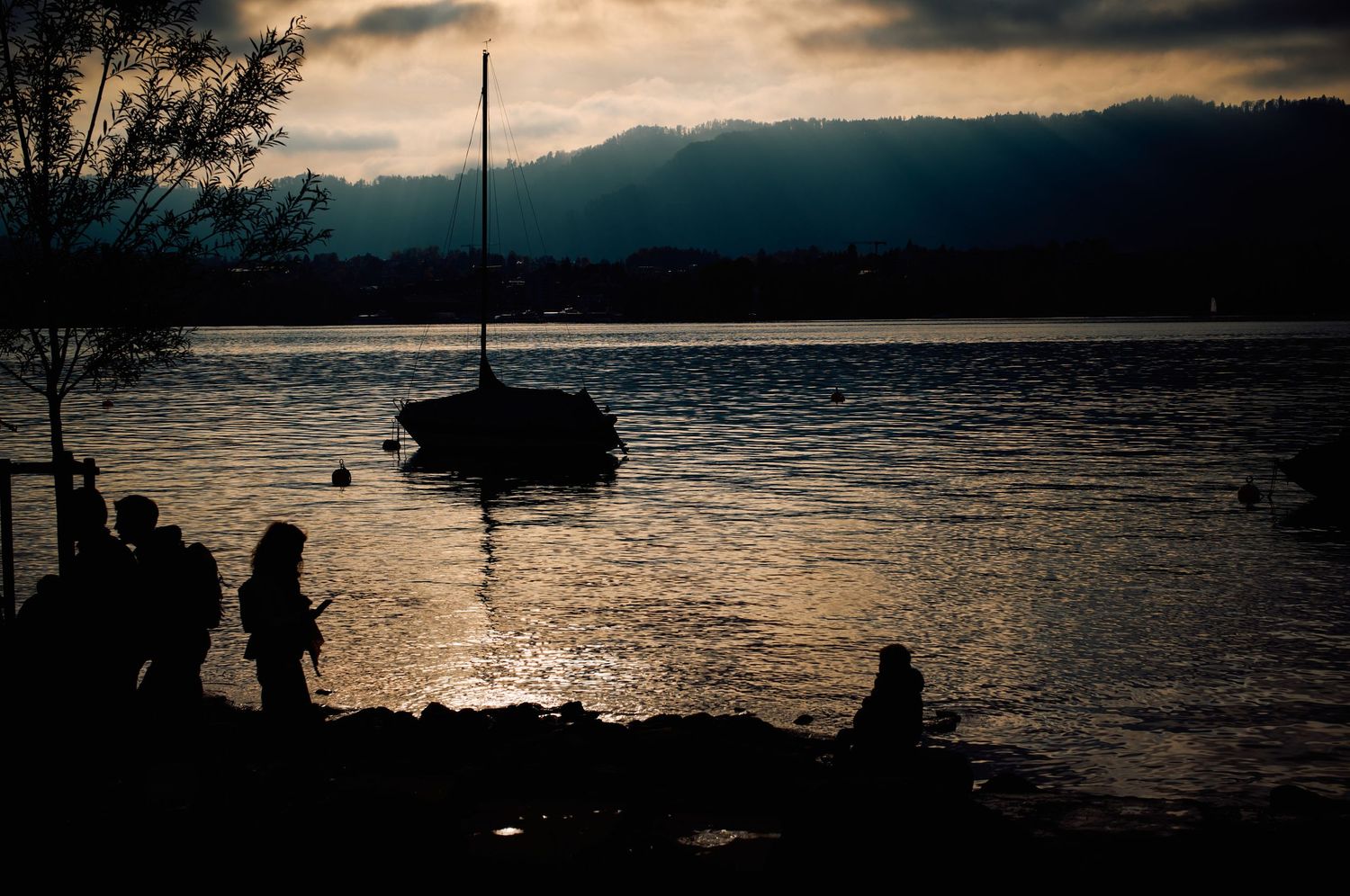 Silhouettes of people watching a boat at sunset on a tranquil lake.