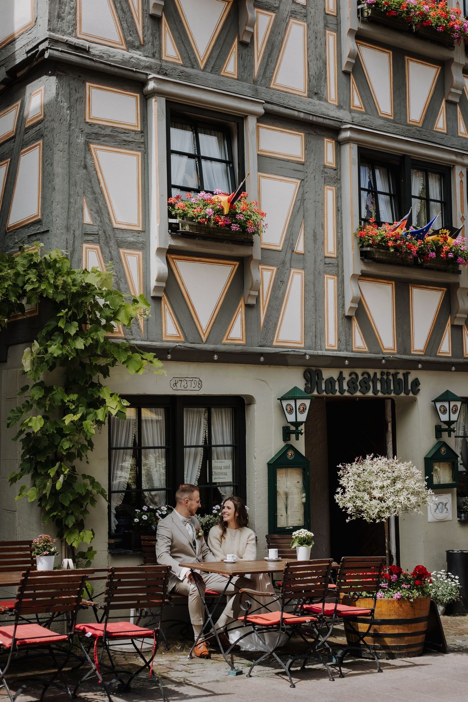 A couple sits at a charming outdoor cafe table in front of a traditional German timber-frame building.
