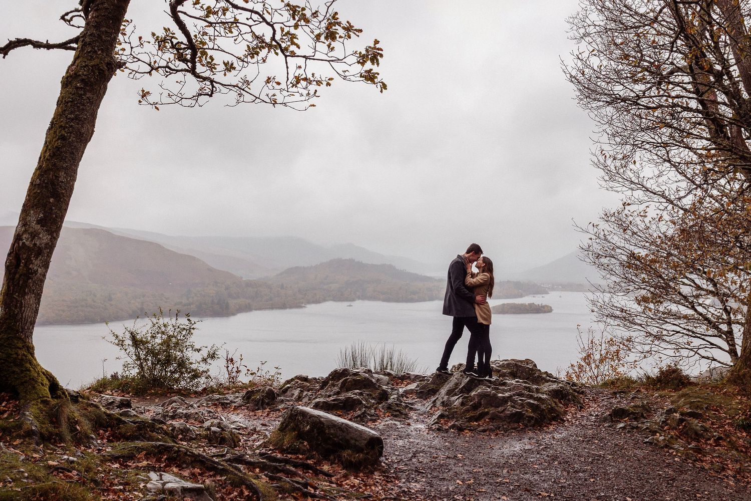 Groom kisses bride's head while she smiles - the backdrop is a country scene with cattle and it's raining