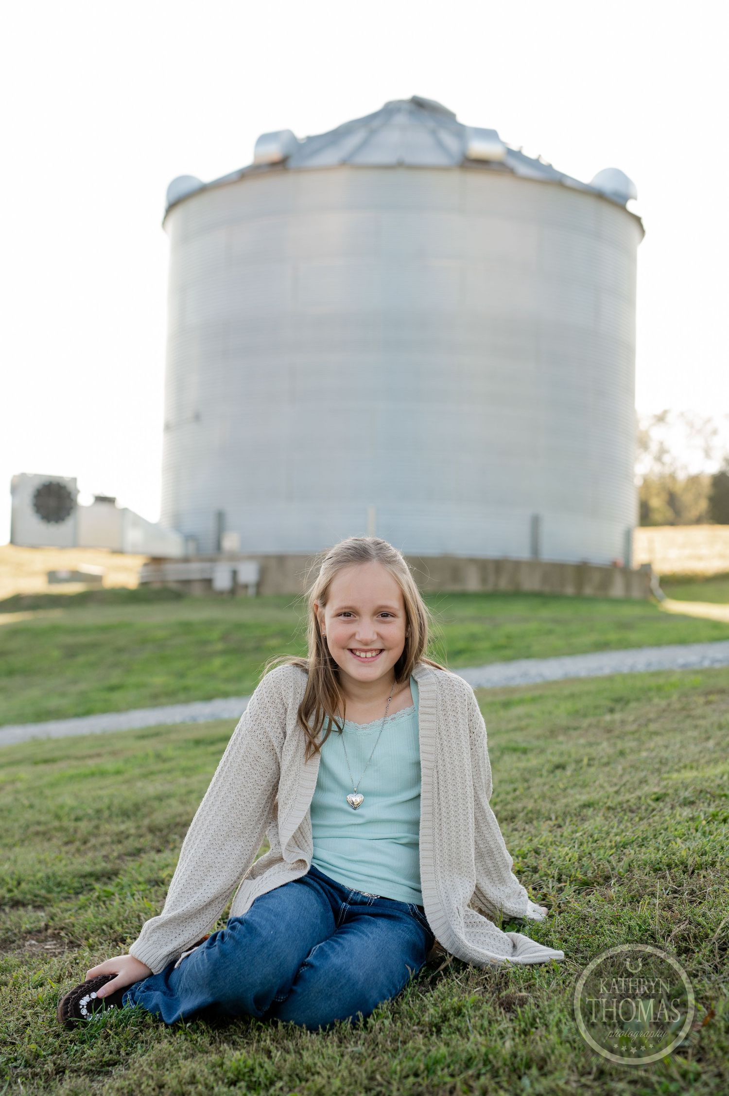 Someone sits casually on grass wearing a cream cardigan and jeans near a towering grain silo.