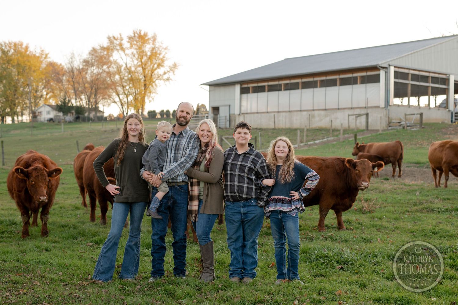 A group poses with Highland cattle grazing in a pasture near farm buildings at sunset.
