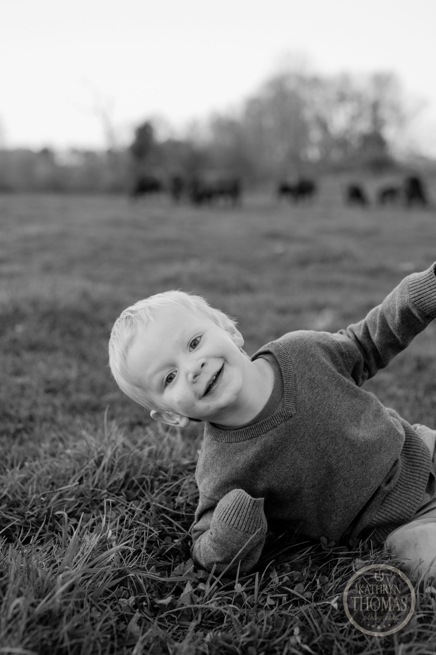 A black and white candid portrait of a playful child with cattle grazing in the distance.