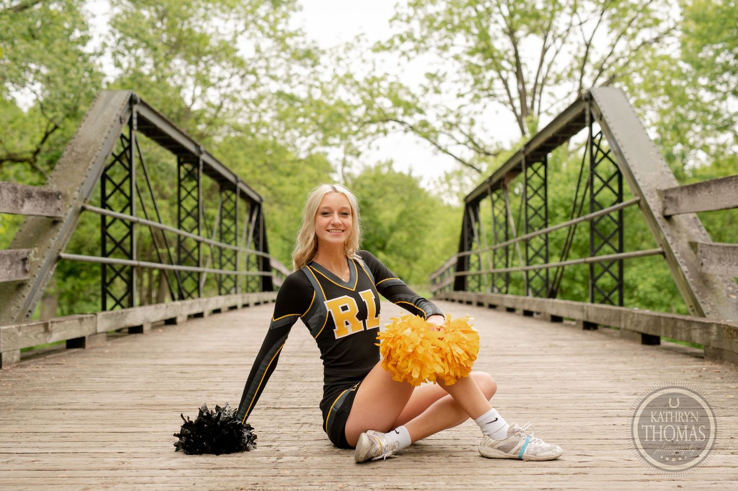 Cheerleader in black and gold uniform posing on wooden bridge with pom poms.