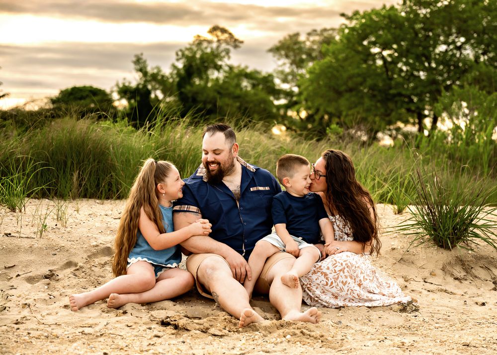A family sits together on a sandy beach at sunset with green foliage in the background.