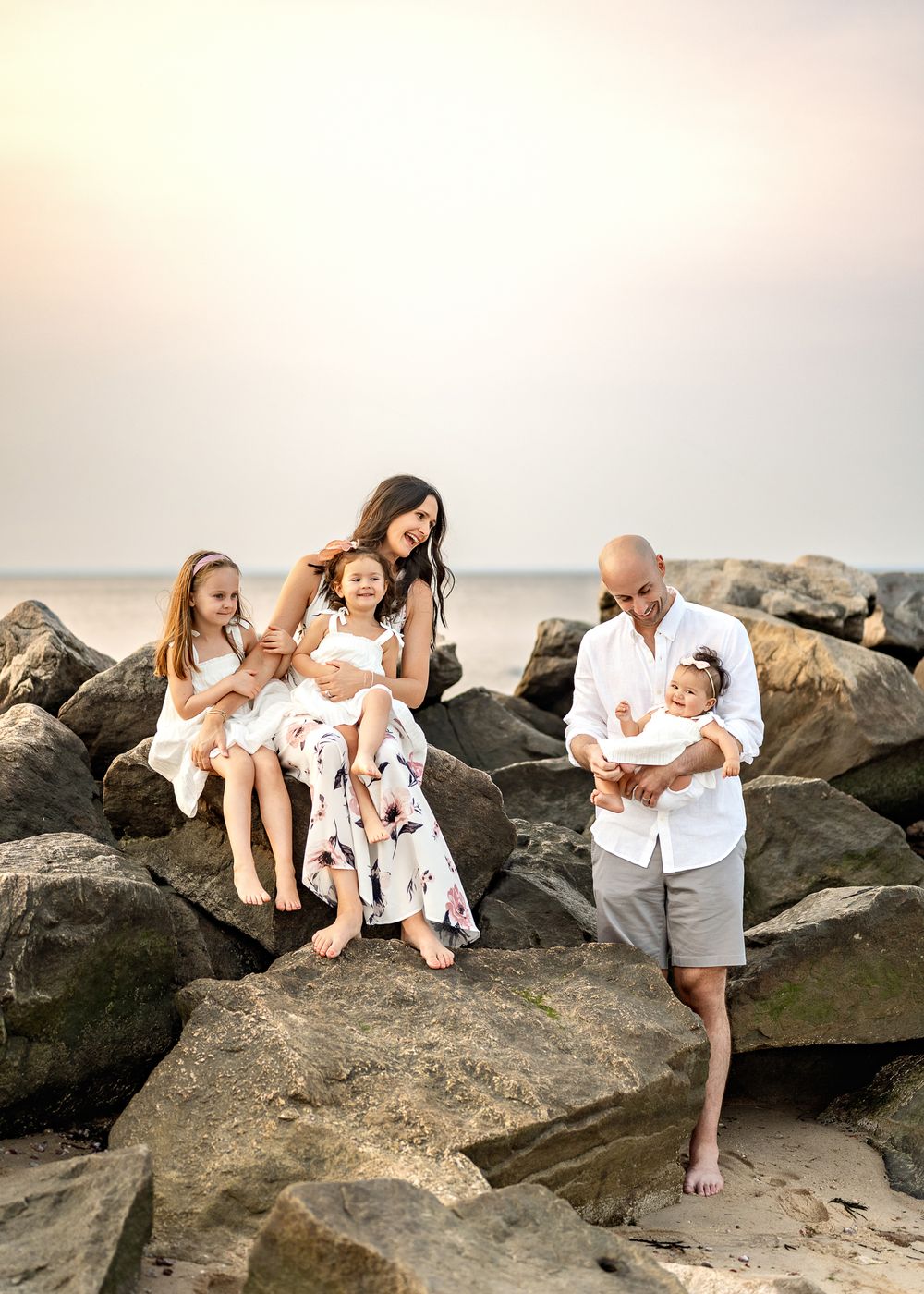 A family in white outfits shares tender moments together while sitting on coastal rocks during sunset.