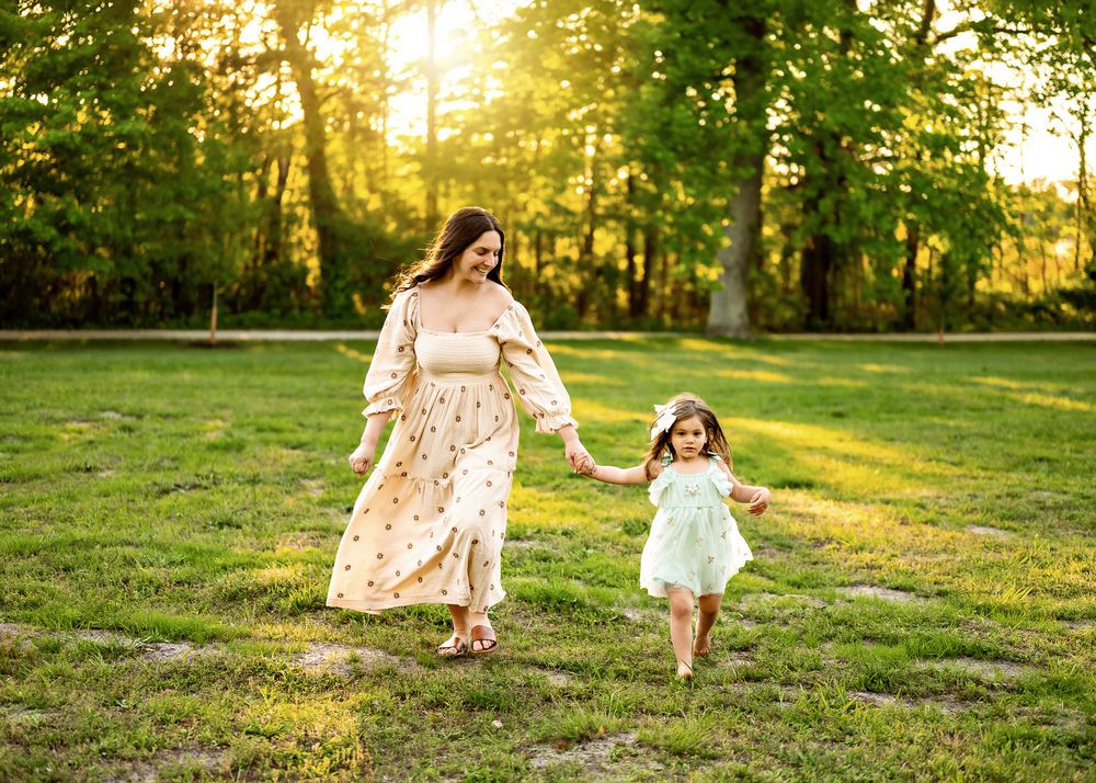 Two figures in flowing dresses hold hands while walking through a sunlit grassy field at golden hour.