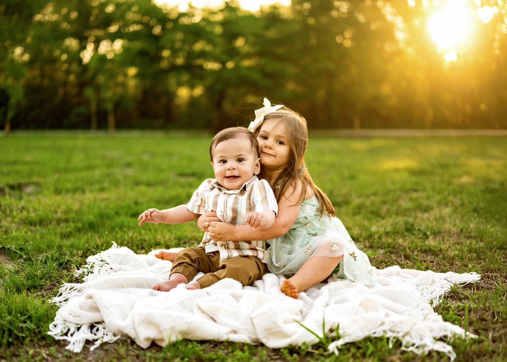 Two children sitting on white blanket in grassy field at sunset with golden sunlight filtering through trees.