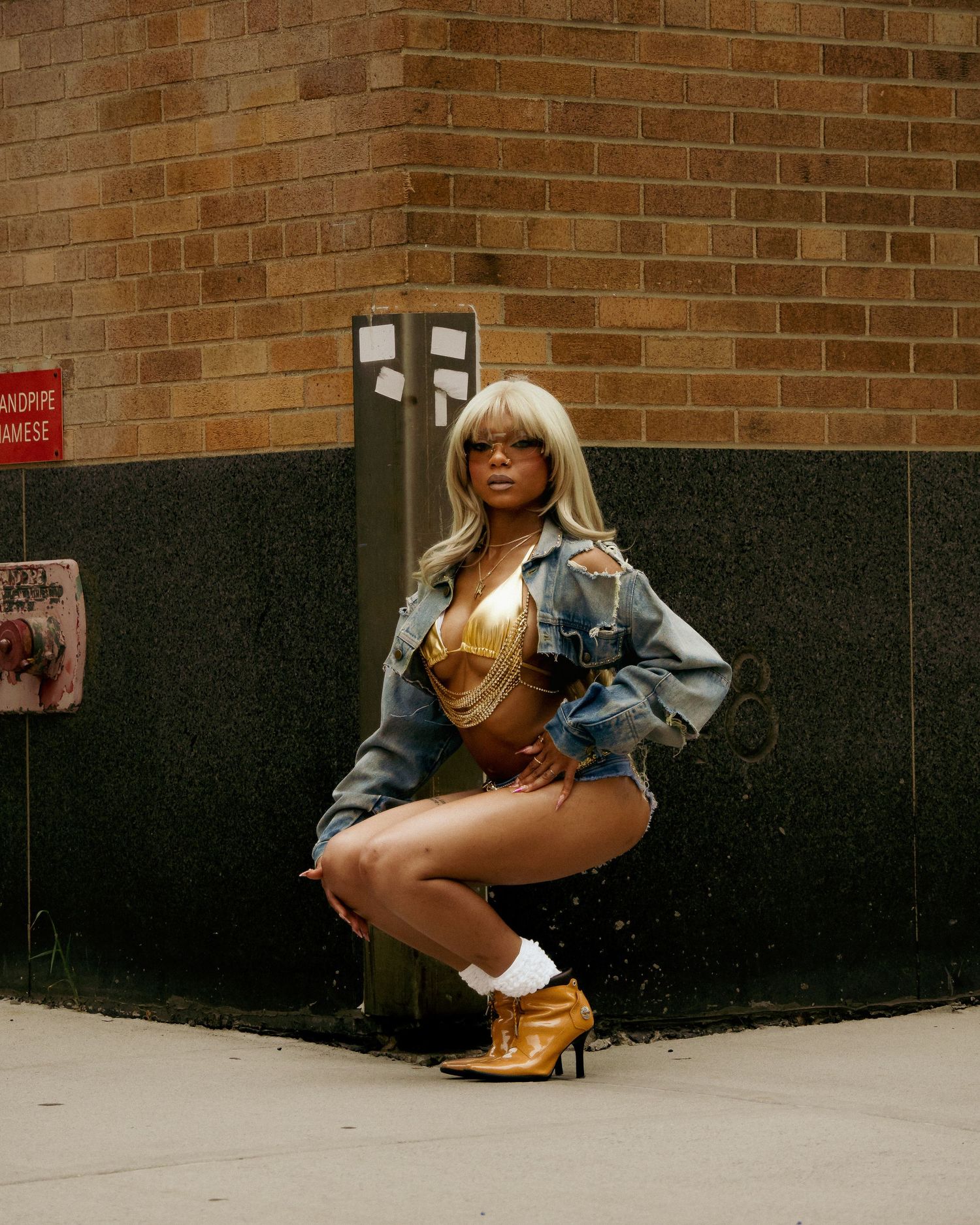 Fashion model posing against brick wall wearing denim jacket and gold boots in urban setting.