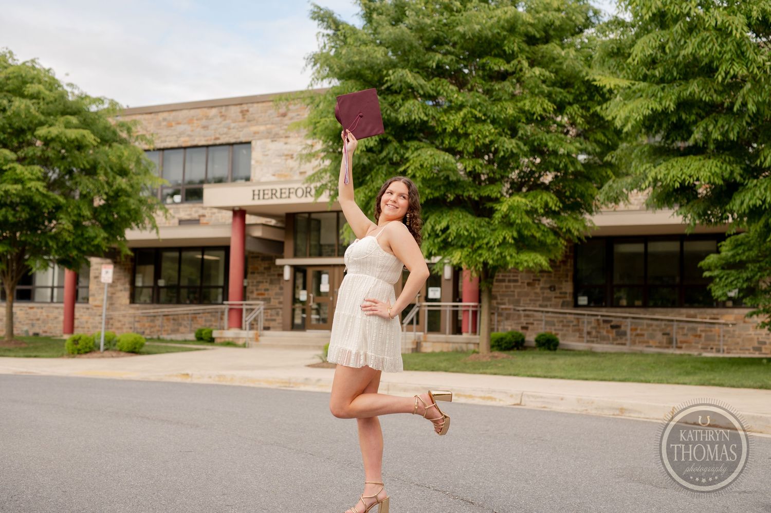 Graduate in white dress strikes celebratory pose while holding burgundy cap outside Hereford High School.