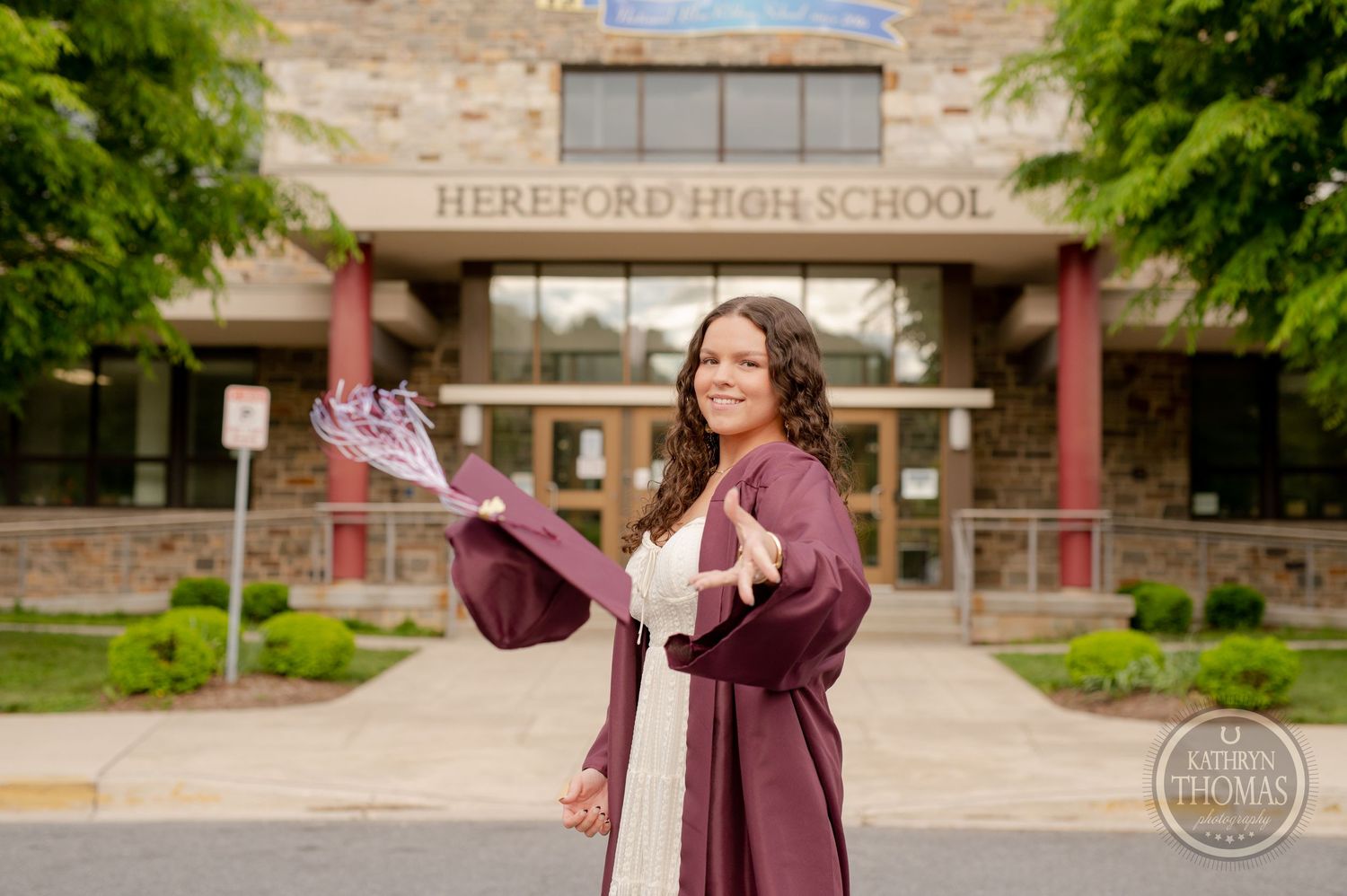 Graduate in maroon gown holds diploma while standing in front of Hereford High School.
