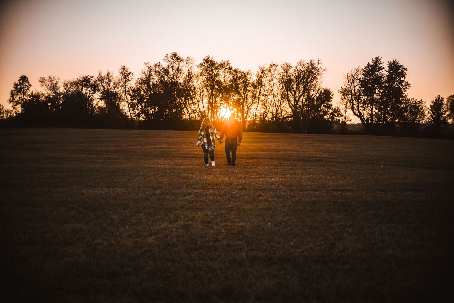 A silhouetted figure stands in an open field at sunset with golden light streaming through trees on the horizon.