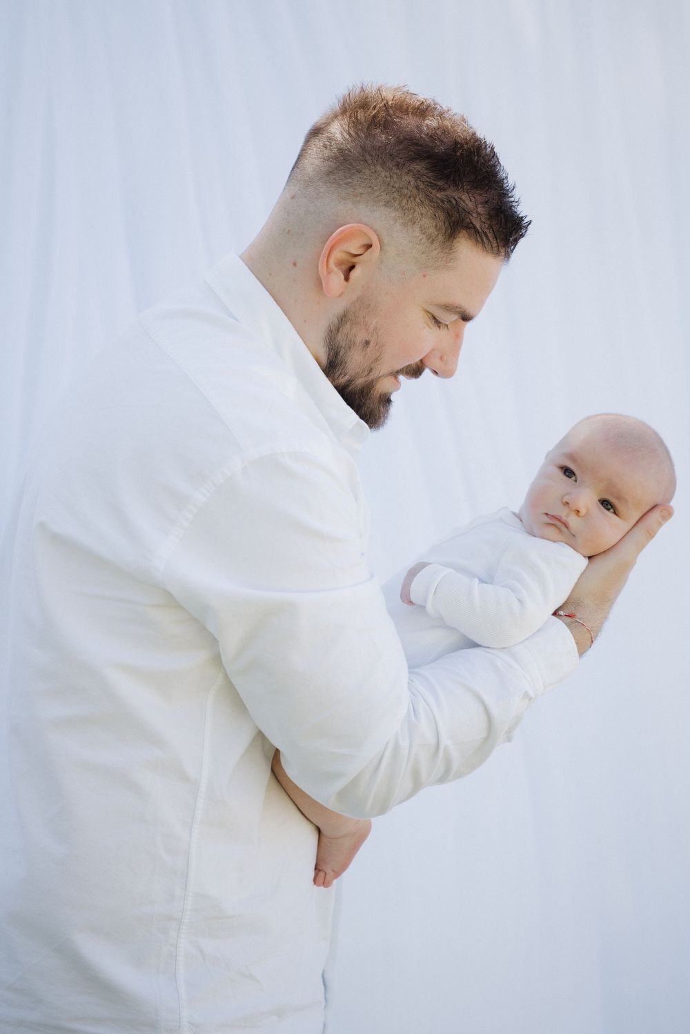 A tender moment between an adult in white clothing holding a small baby dressed in light colored clothing against a light background. locati
