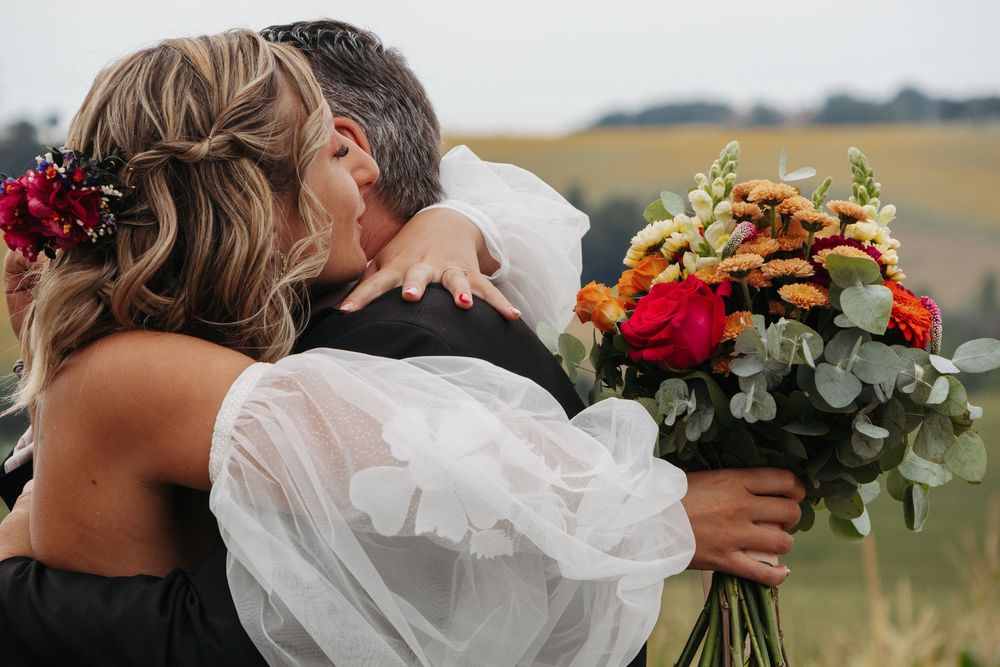 Bride embracing groom holding vibrant red and orange wedding bouquet in field.