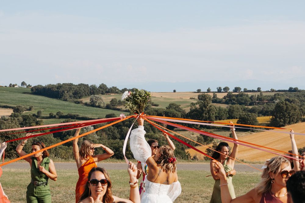 Bride tossing bouquet with bridesmaids holding orange ribbons on scenic hilltop.