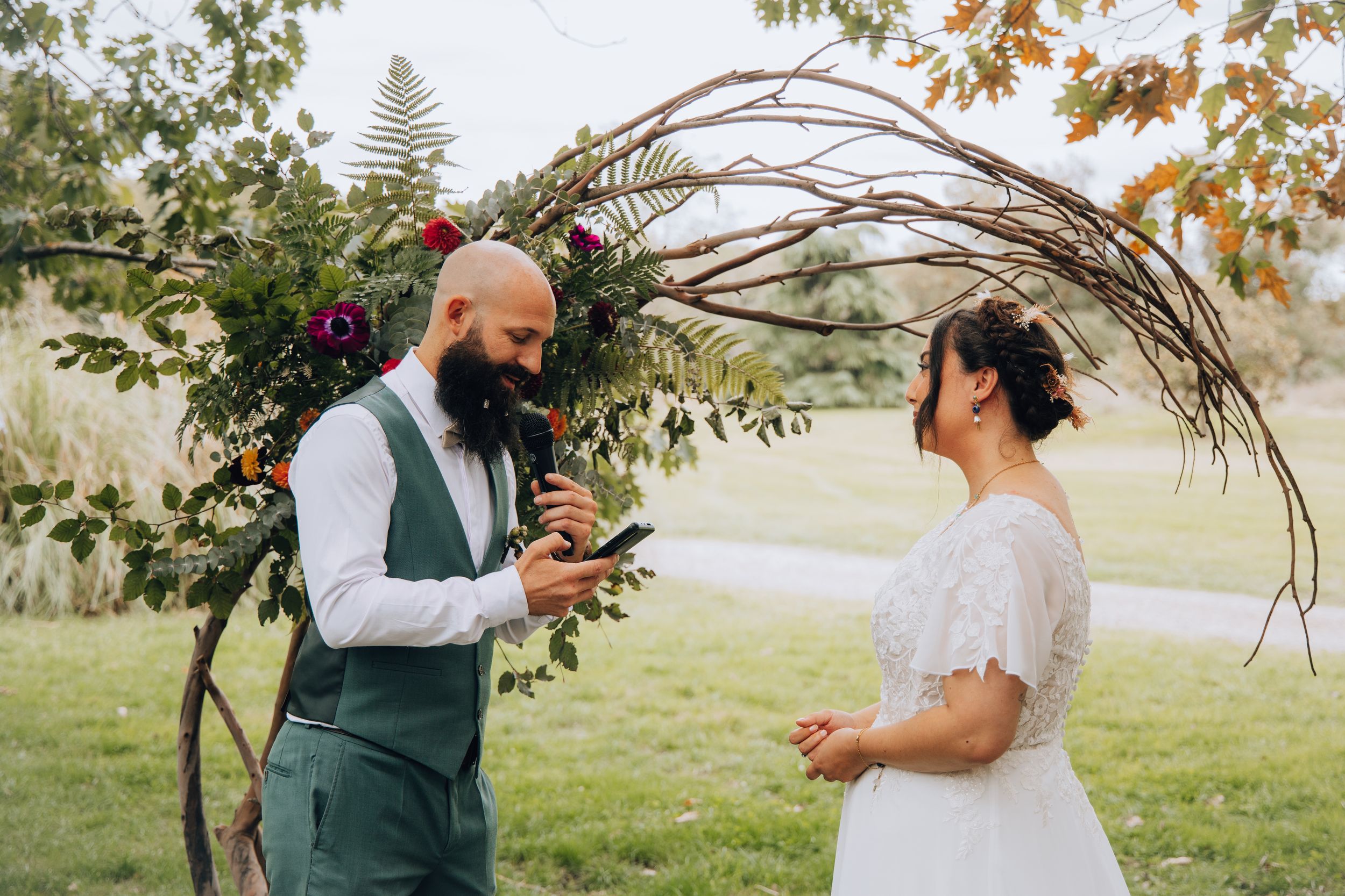 Groom reading wedding vows to bride under a floral arch during outdoor ceremony.