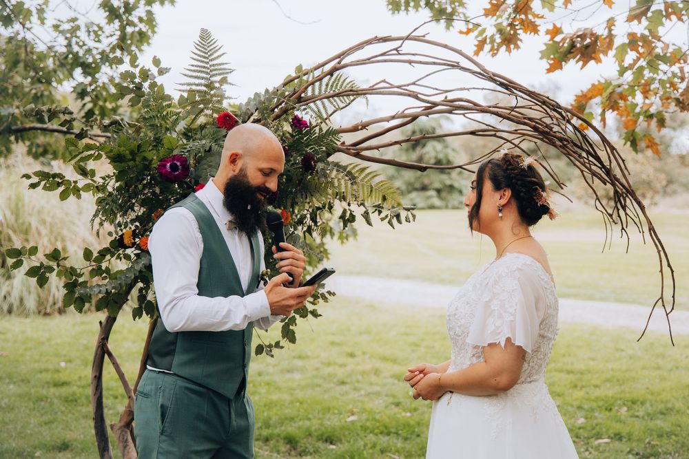 Groom reading wedding vows to bride under a floral arch during outdoor ceremony.