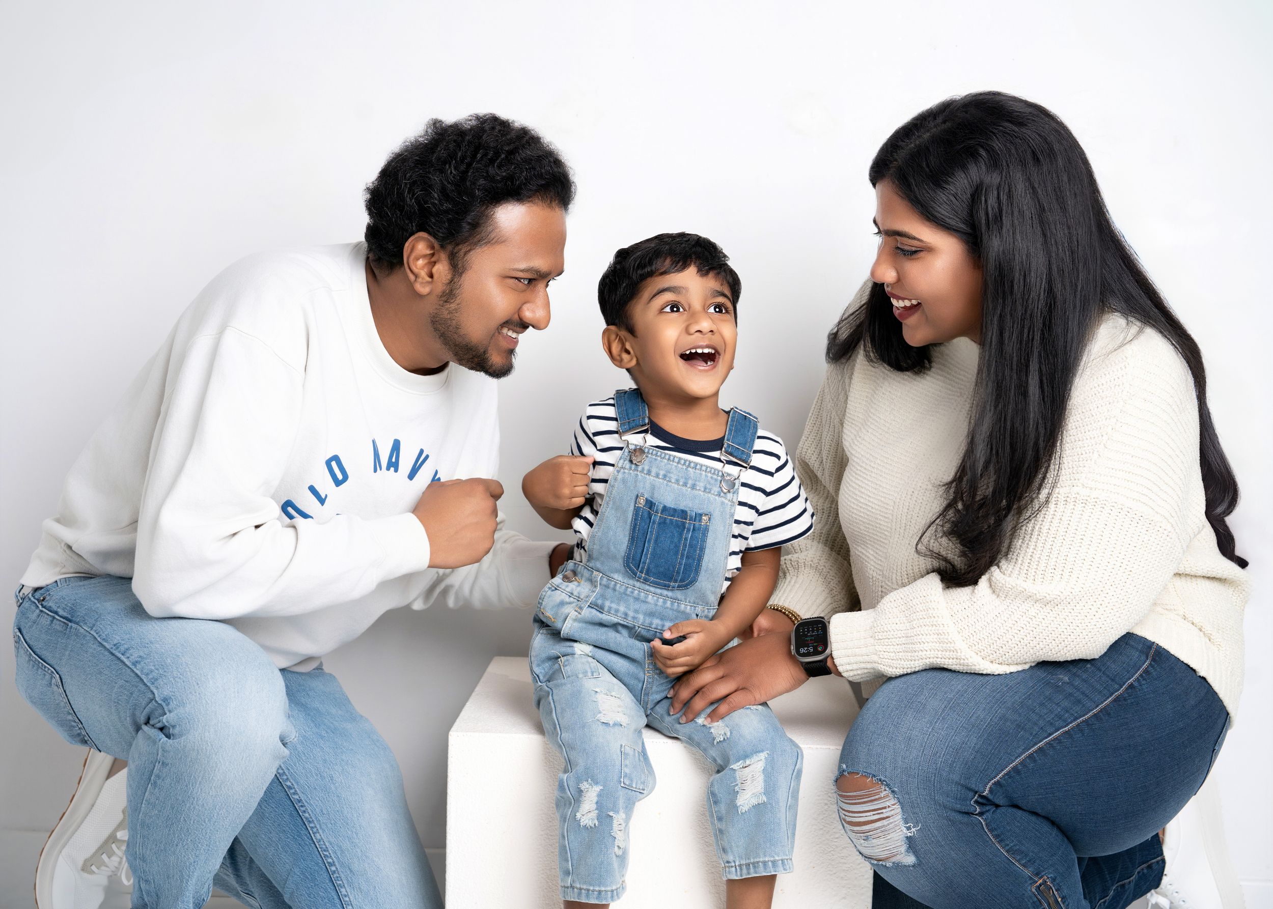 A family wearing casual clothes sits together on a white backdrop, laughing and sharing a joyful moment.