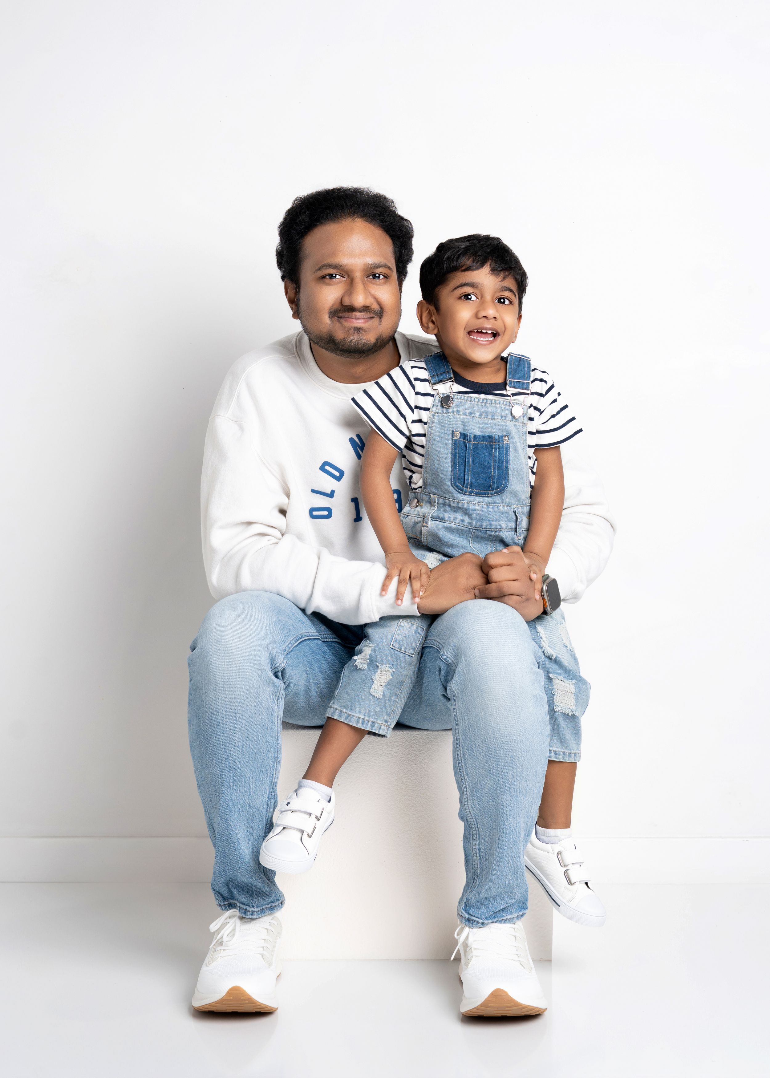 An adult and child wearing denim and white outfits sit together on a white stool against a light background.