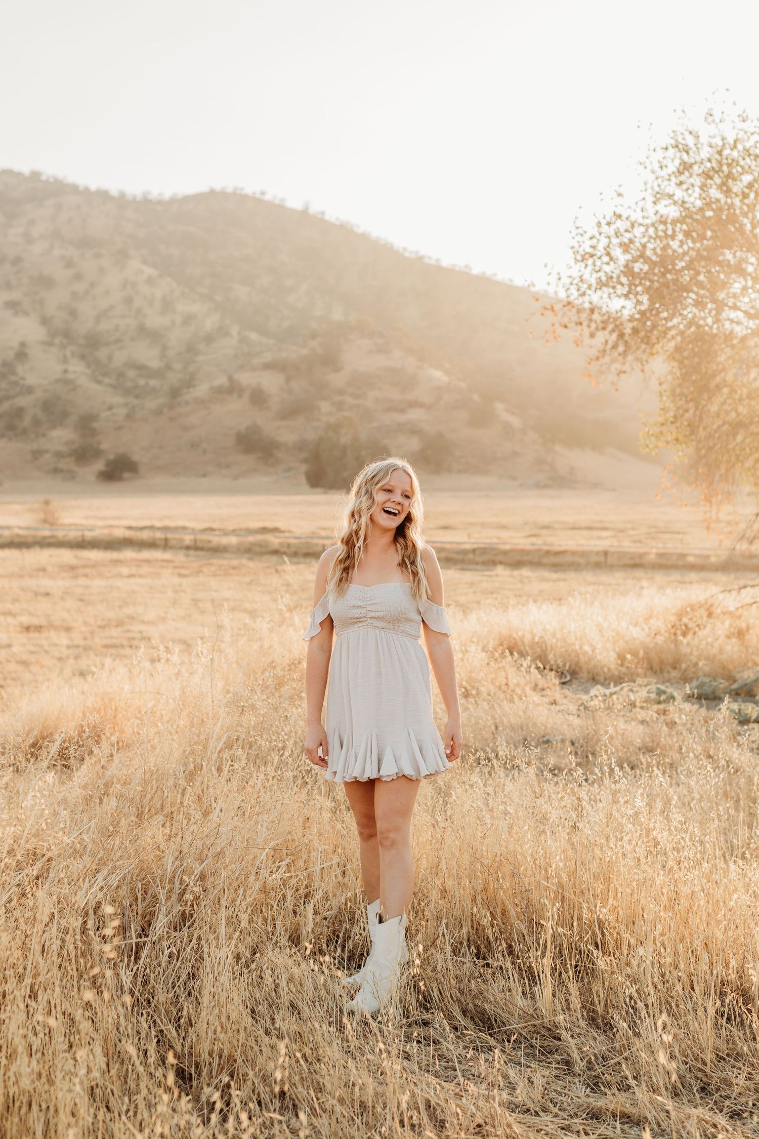 Person in light dress walking through golden sunlit field with mountains in background.