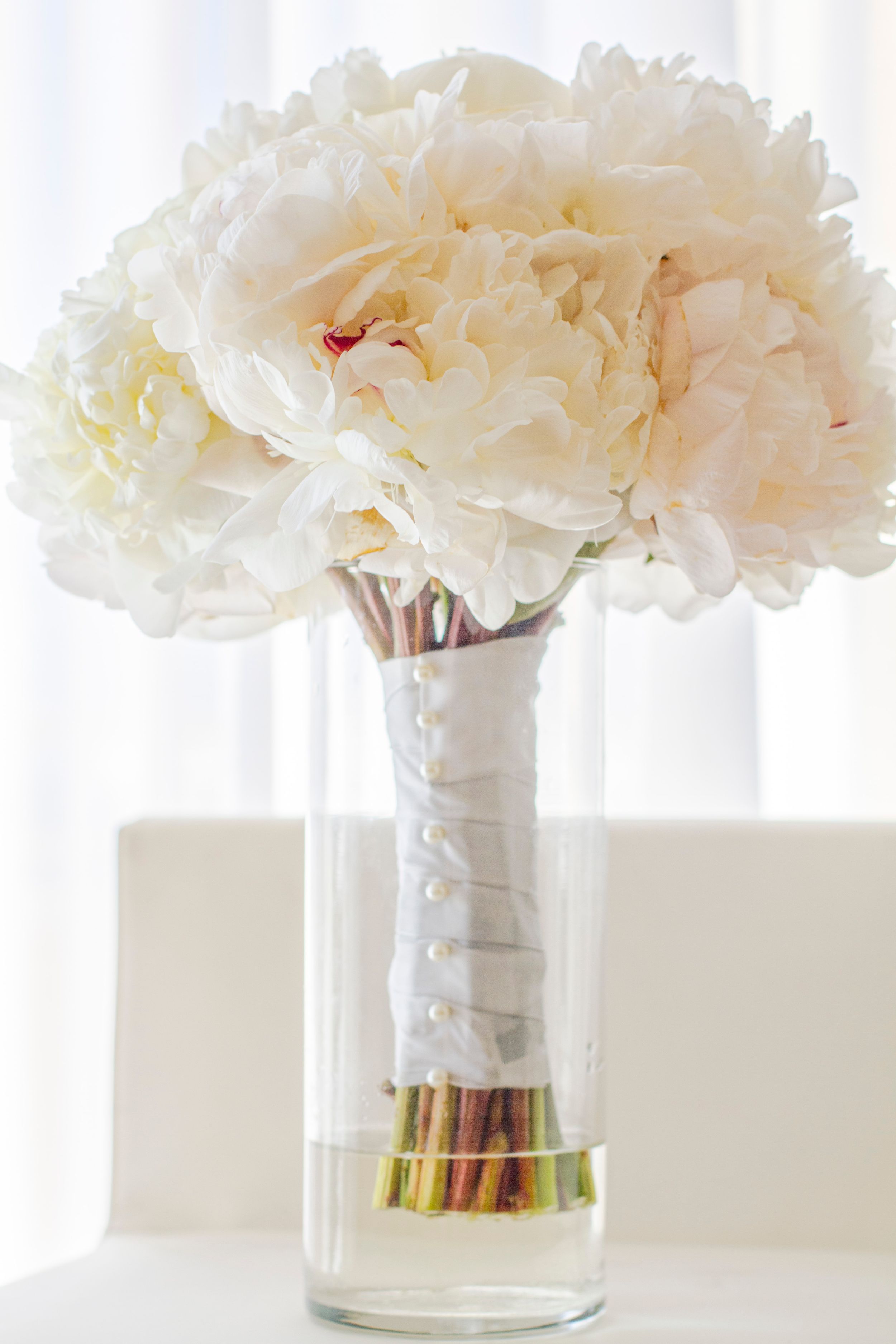 White peony bridal bouquet wrapped in white ribbon with pearl pins displayed in glass vase.