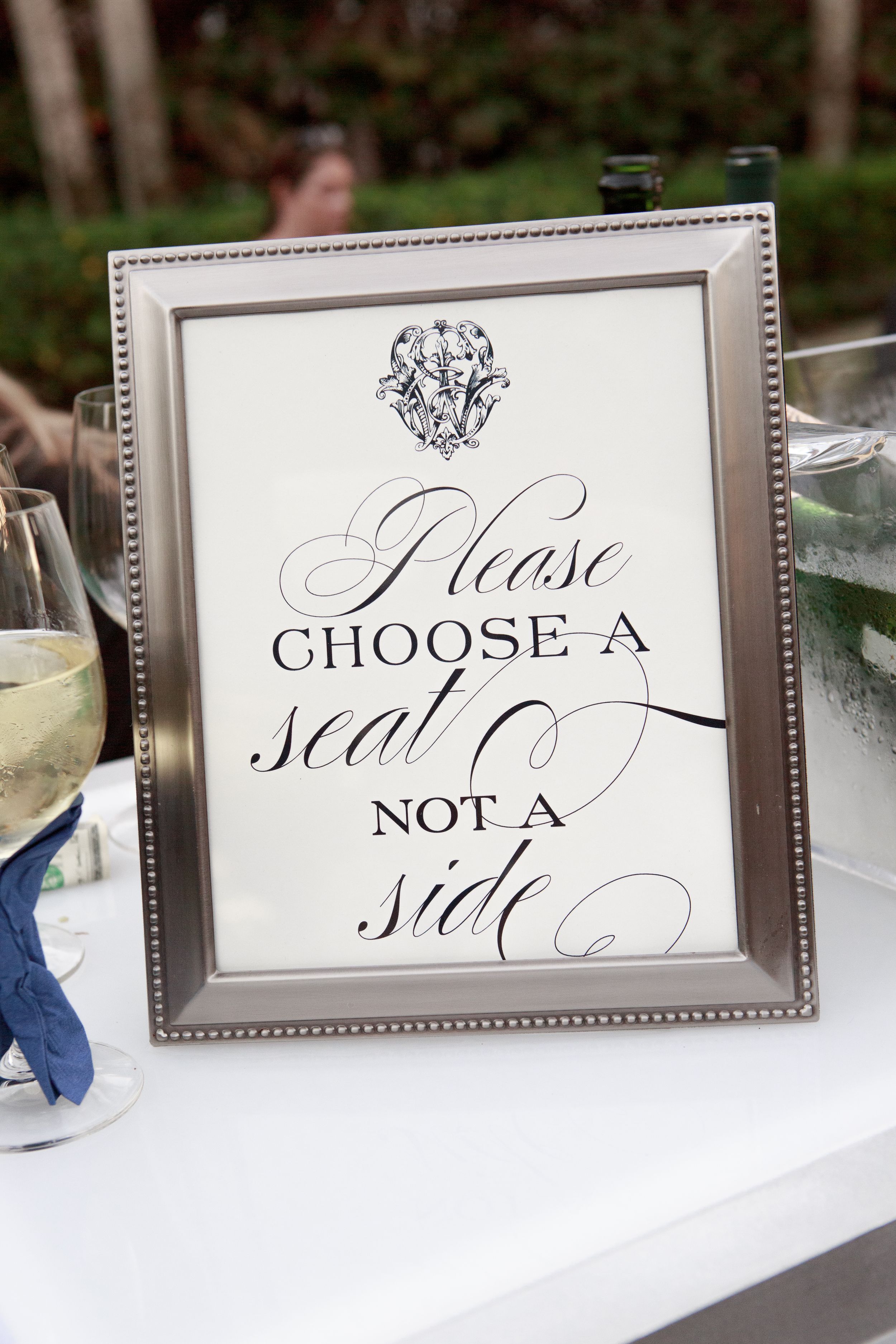 Silver framed wedding sign reading Please Choose a Seat Not a Side in elegant calligraphy. Naples