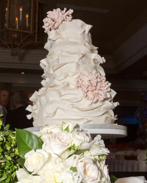 Elegant white tiered wedding cake with ruffled fondant and pink peony flowers surrounded by white roses.