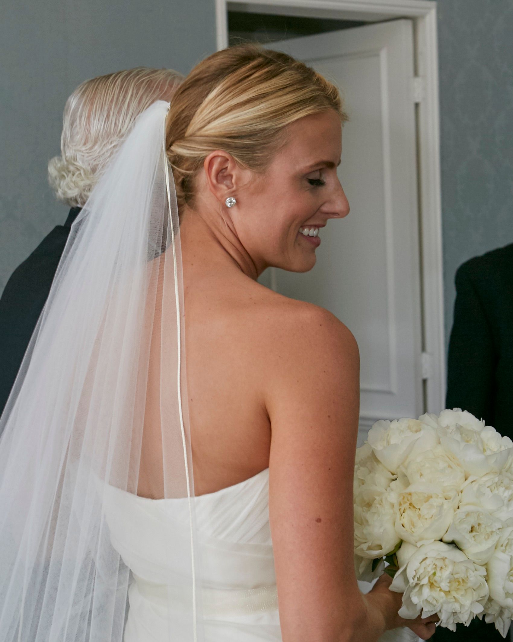 Blonde bride in strapless white gown with veil and updo hairstyle holding white peony bouquet. Naples
