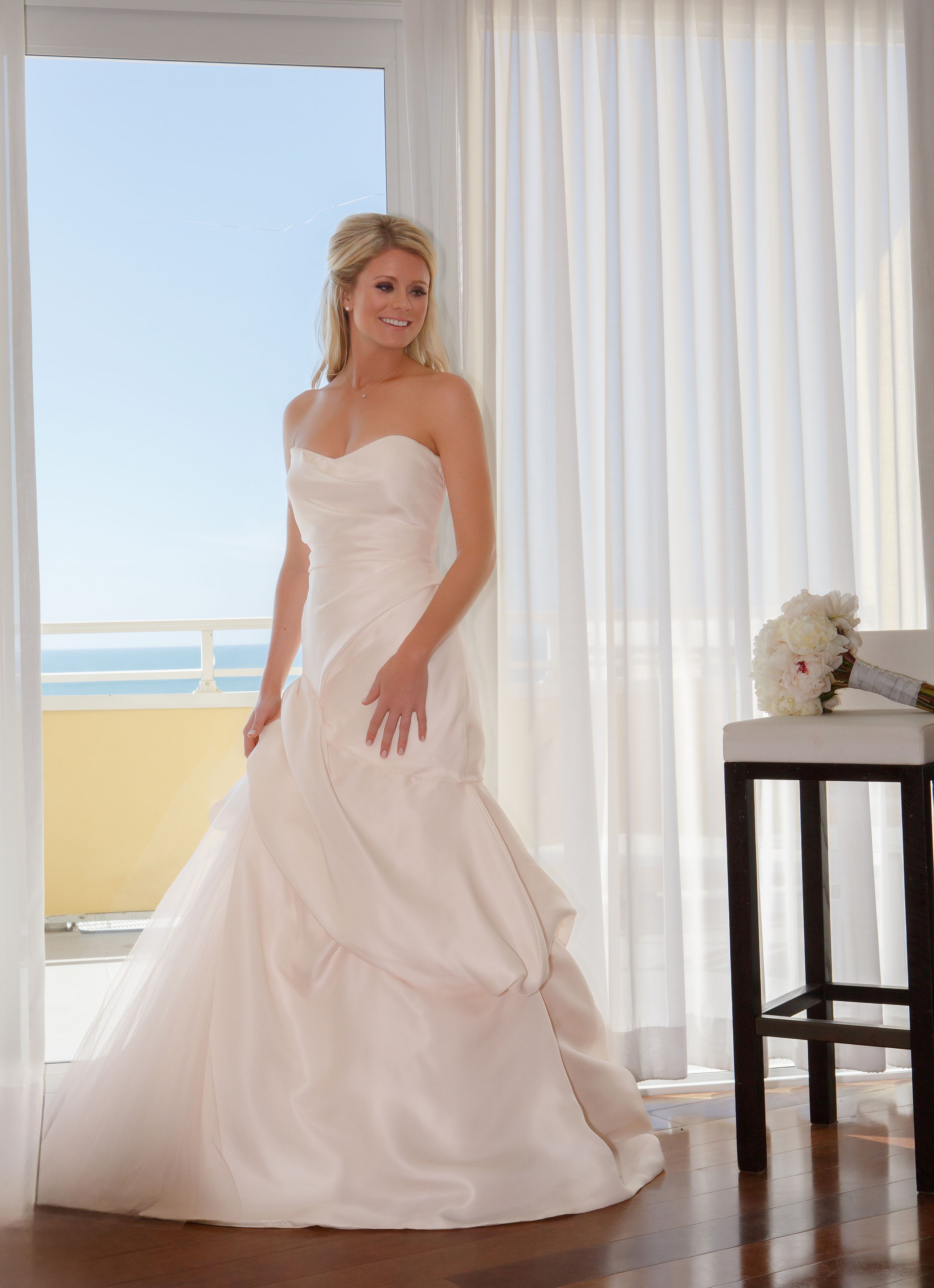 Radiant bride in strapless ball gown wedding dress standing by ocean-view window. Naples