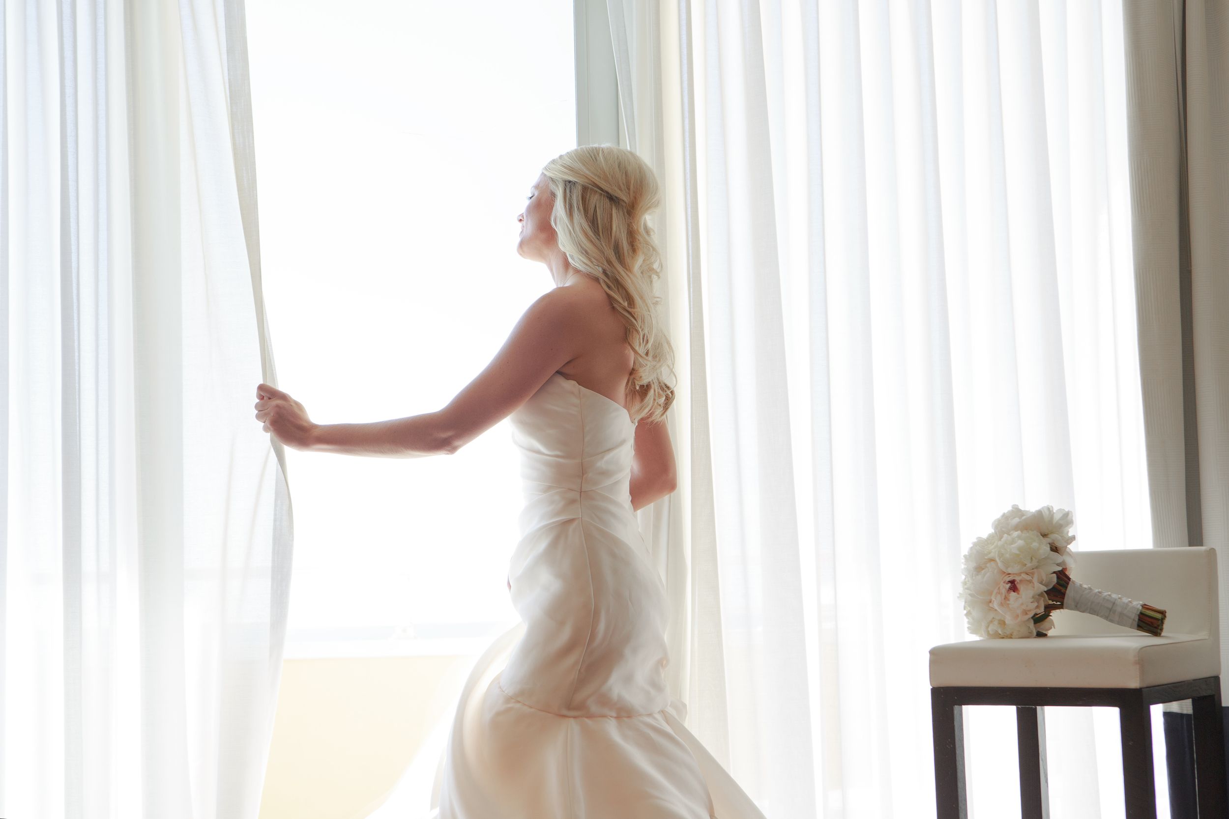 Elegant blonde bride in strapless white wedding gown gazing out large curtained window. Naples