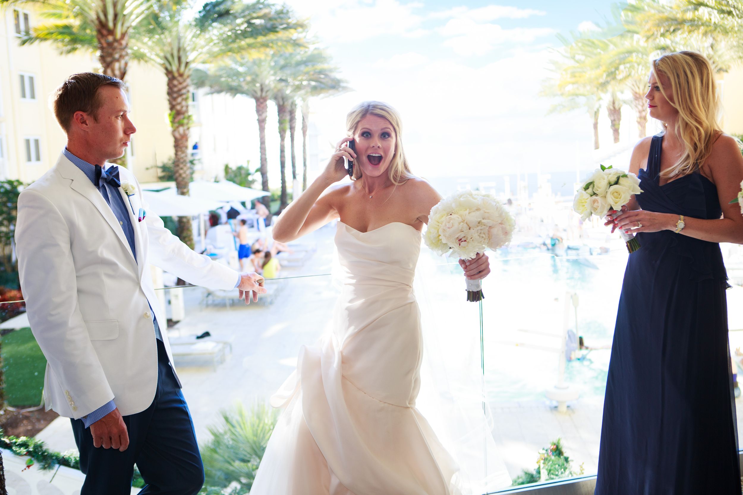 Bride in strapless white wedding gown laughing on phone with bridesmaid in navy dress at outdoor venue.