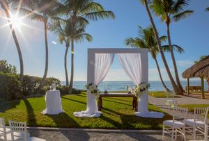 Beachfront wedding ceremony setup with white draped arch, palm trees, and ocean view at sunset. Naples