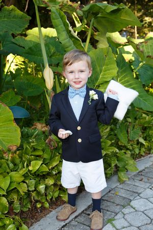 Adorable ring bearer in navy blazer and bow tie holding pillow in garden setting.