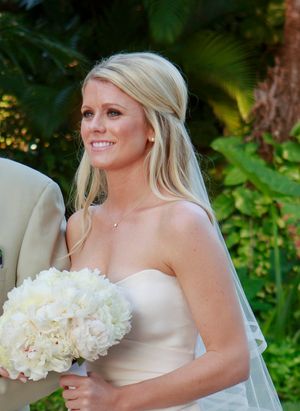Smiling blonde bride in strapless ivory gown holding white peony bouquet outdoors among tropical palms. Naples