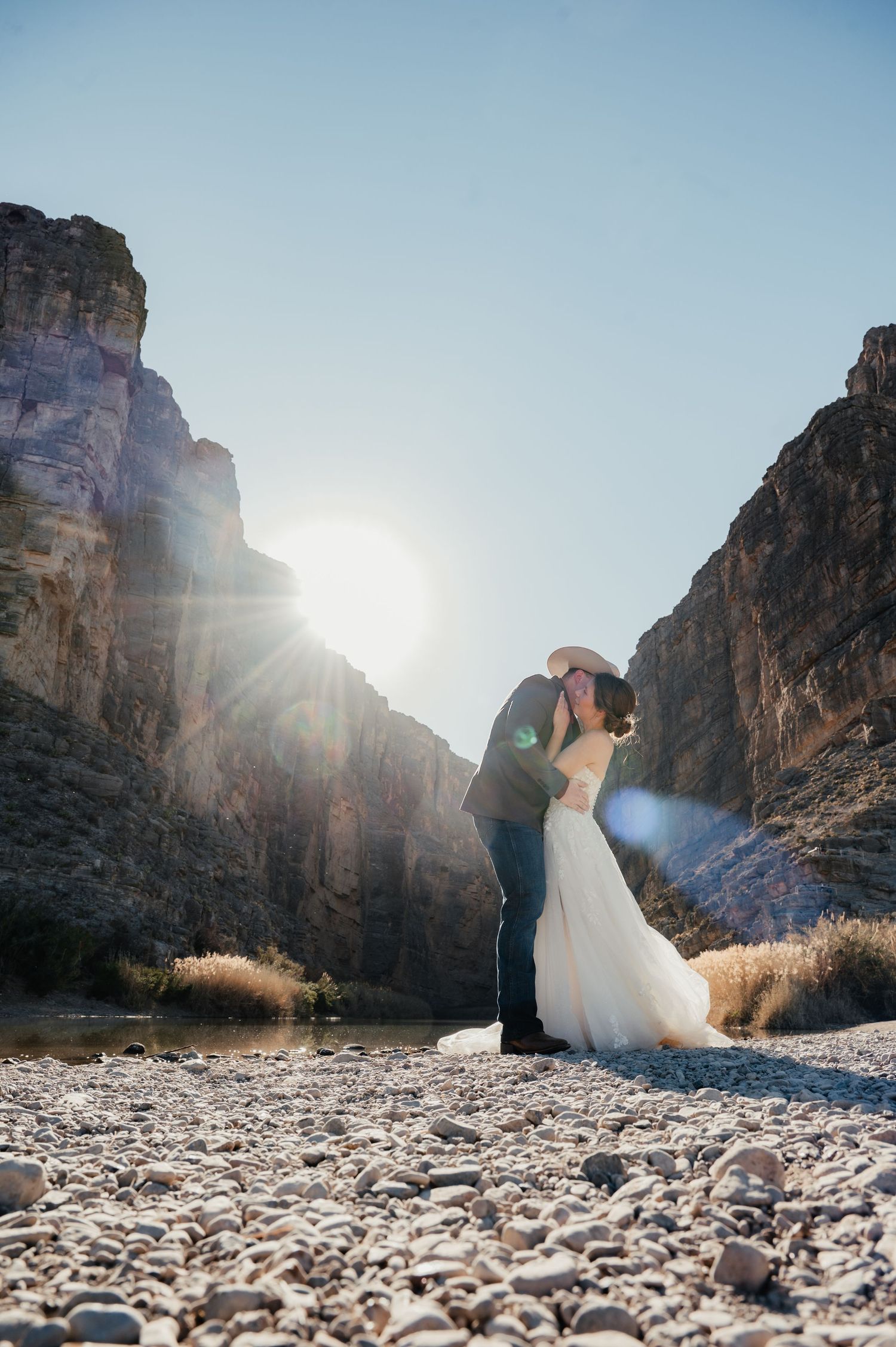 Two people share an intimate moment along a rocky riverbed surrounded by towering canyon walls as sunlight streams through.