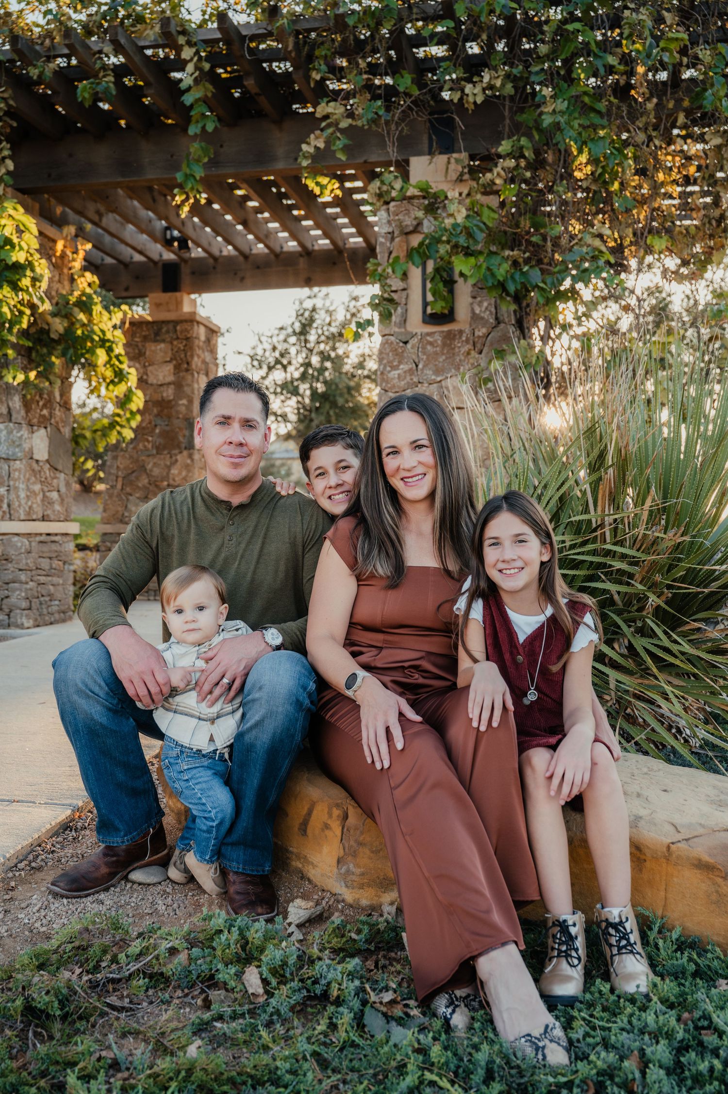 A family sits together outdoors under a pergola with climbing vines in a garden setting during golden hour.