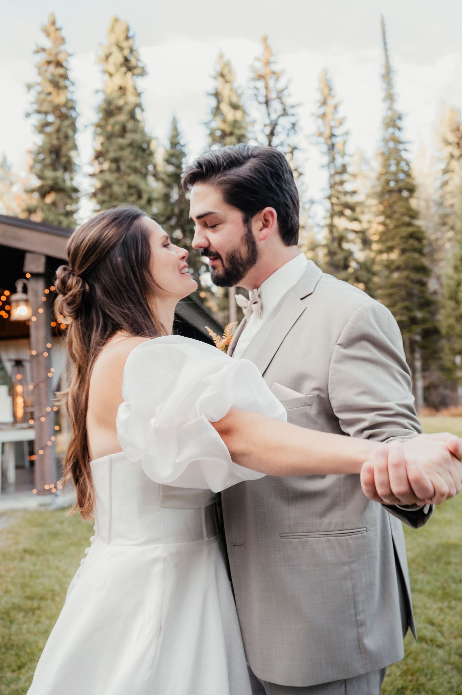 A couple shares their first dance outdoors with tall evergreen trees and string lights in the background.
