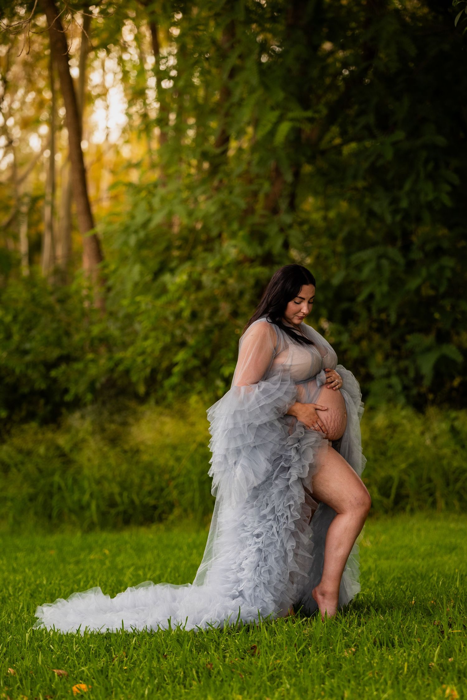 A maternity photoshoot featuring a flowing gray tulle gown in a sunlit forest setting.