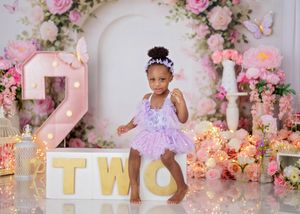 Toddler girl in lavender tutu dances beside illuminated TWO letters with pink floral backdrop.