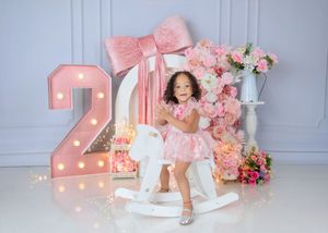 Toddler girl in pink dress rides rocking horse beside illuminated number 2 with floral backdrop.