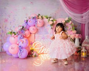 Baby girl in white pink dress stands by floral arch with pink balloons and neon 'One Mrs' sign.